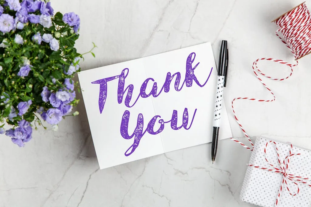 A stylish thank you card paired with purple flowers, a gift box, and pen on a marble surface.
