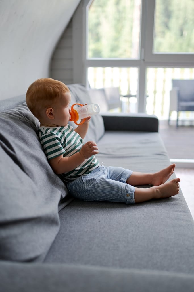 Cute baby in striped shirt relaxing on a sofa, drinking from a bottle in a cozy home setting.
