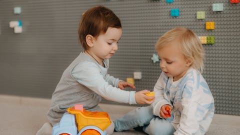 Two toddlers happily playing with a toy truck indoors, surrounded by colorful blocks.