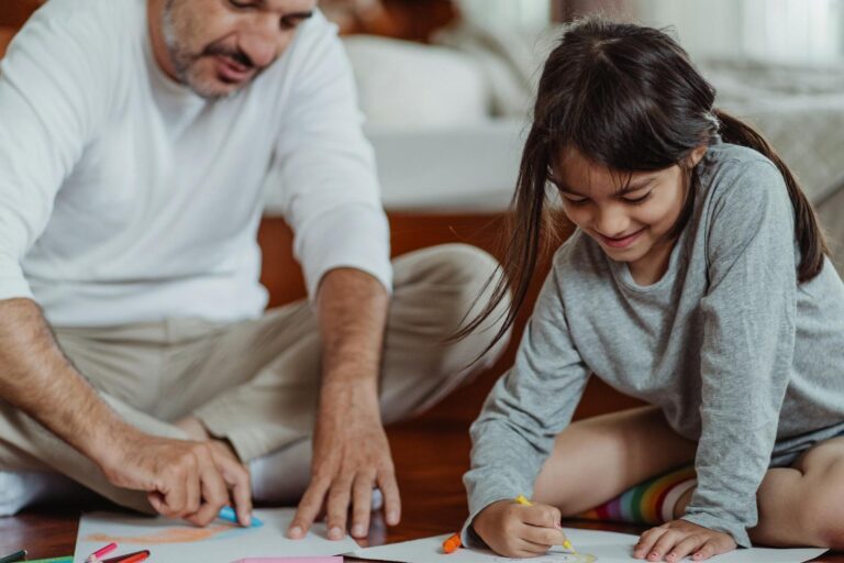 Smiling father and daughter bonding over art and crafts at home, drawing with colorful crayons.
