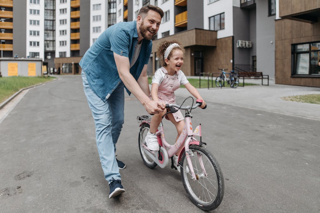 A joyful moment as a father helps his daughter ride a bicycle in an urban setting.